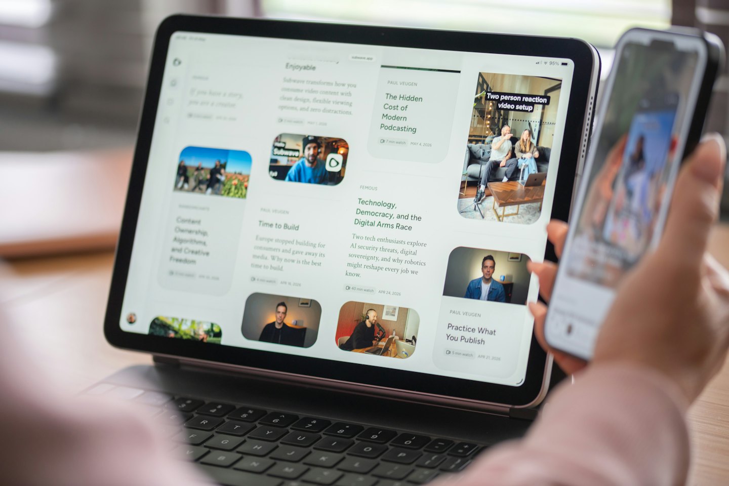 Hands holding a smartphone with a tablet and keyboard.